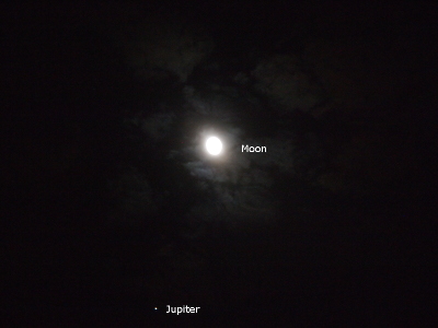 [This sky view has clouds passing by and in front of the moon. The light reflecting from the moon to the clouds lights the sky. Jupiter near the bottom of the image is a bright white dot. White text labeling the moon and Jupiter were added to the right of each orb.]